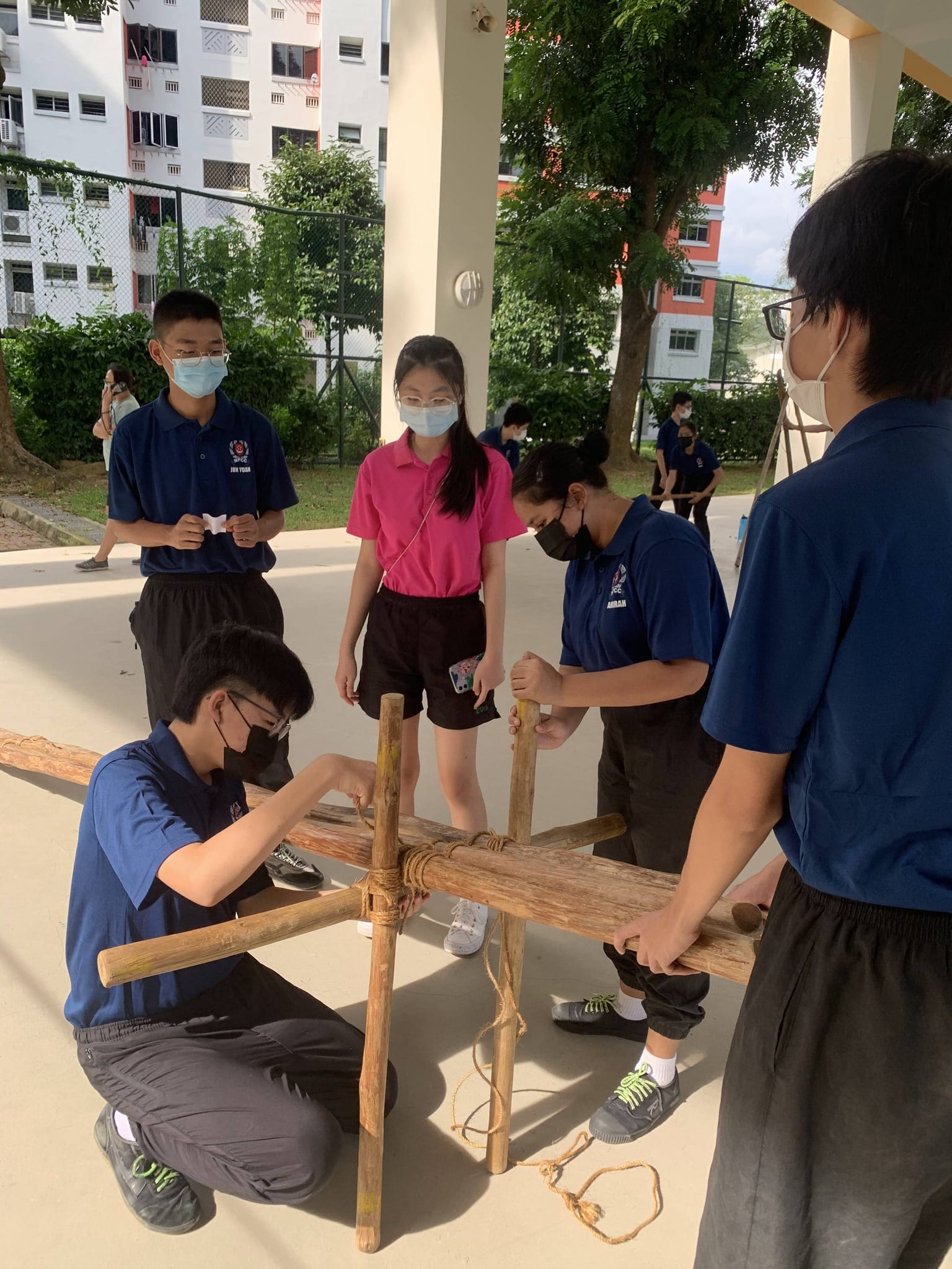 Students wearing masks work together outdoors to assemble a wooden structure tied with rope on a paved area.